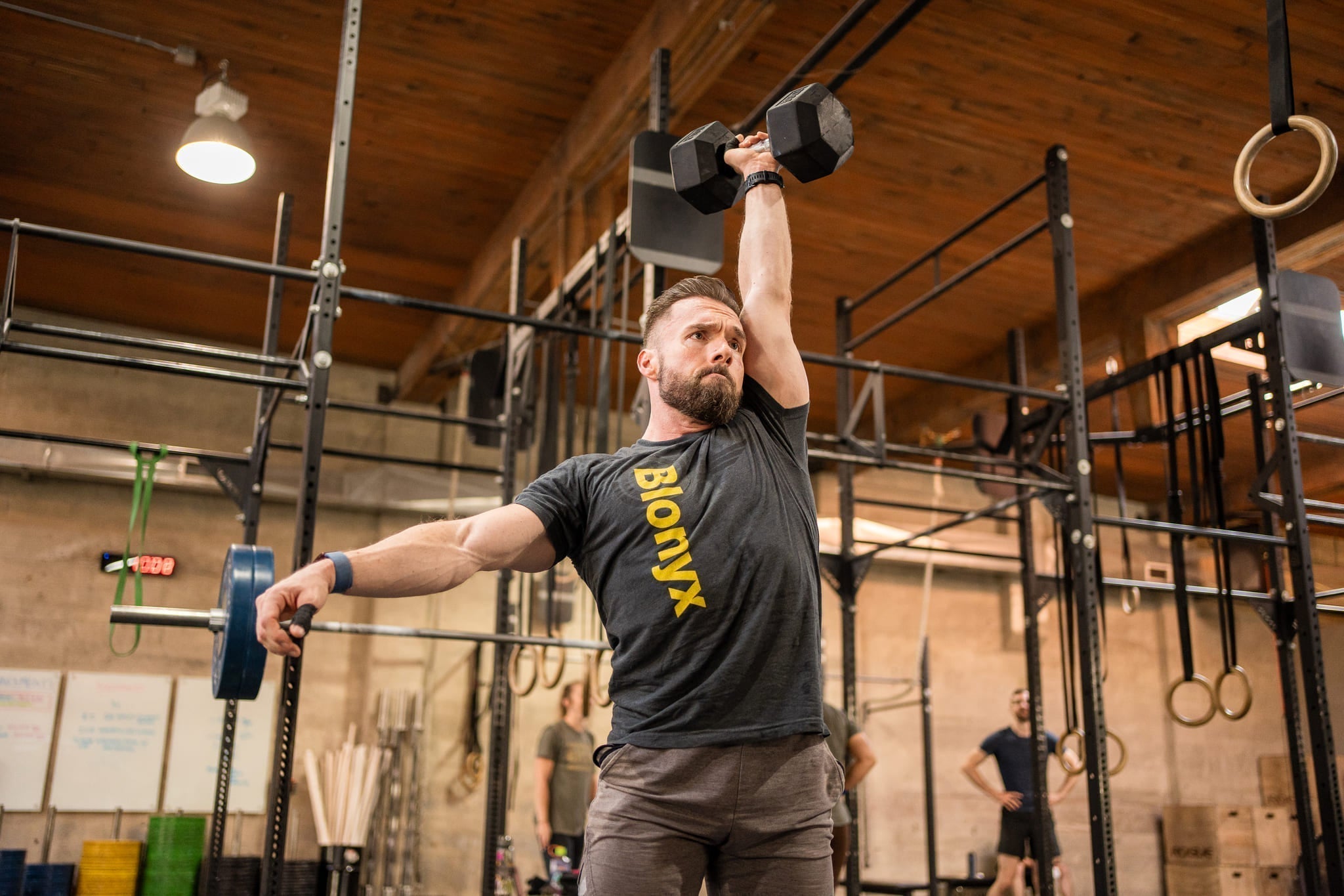Jason Marchand, wearing a Blonyx t-shirt, performs a single-arm dumbbell snatch overhead in a CrossFit-style gym.