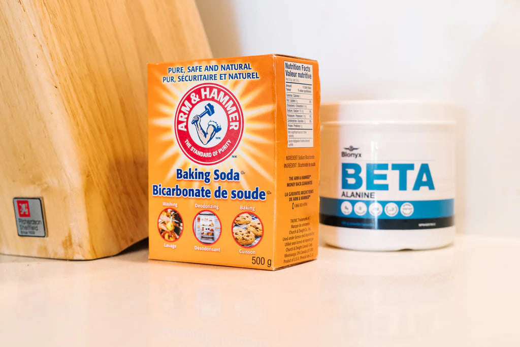 A box of Arm & Hammer Baking Soda next to a white tub of Beta Alanine, set on a light surface with a wooden cutting board in the background.