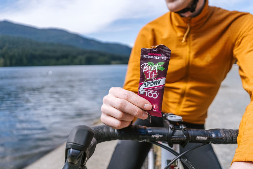 Cyclist holding Beet It Sport Top Up 100 energy gel beside a mountain lake before an endurance ride, providing carbohydrates and dietary nitrates for sustained performance.
