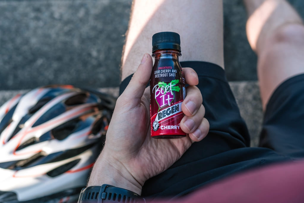 Cyclist holding a Beet It Regen Cherry+ tart cherry recovery shot after a ride, supporting post-exercise recovery and muscle function.
