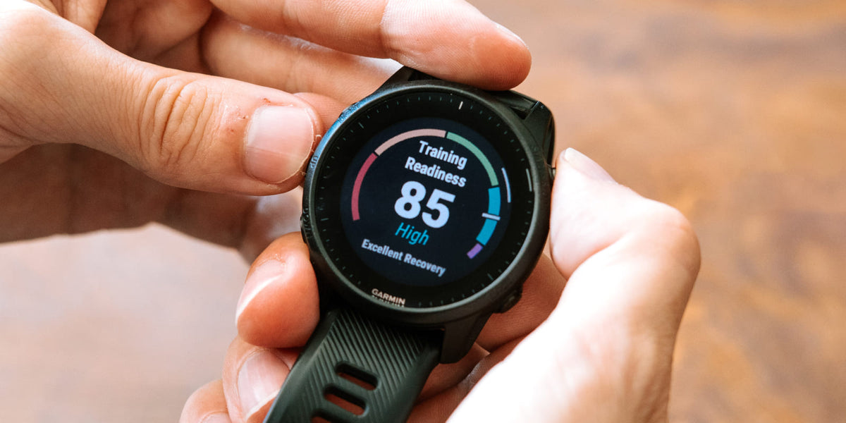 A close-up of a black Garmin smartwatch held in two hands, showing a training readiness screen with 'Training Readiness 85 High' on the display.