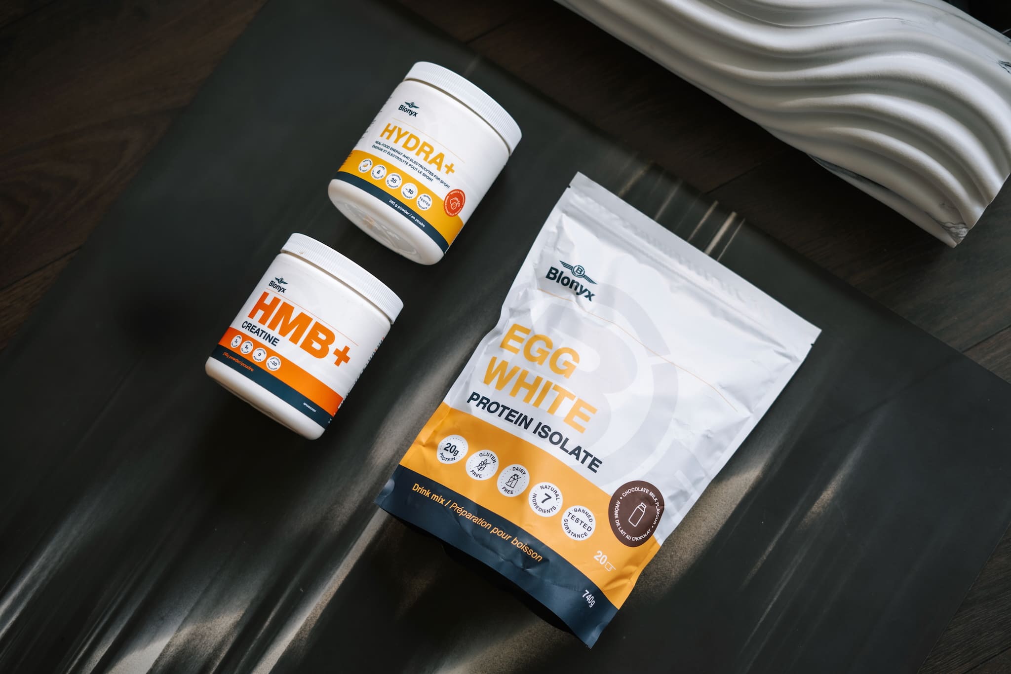 Three Blonyx products (a tub of Hydra+, a tub of HMB+ Creatine, and a bag of Egg White Protein Isolate) on a yoga mat. 