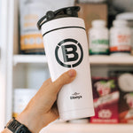 White tumbler with black logo held by a hand in front of a shelf with products.