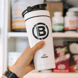 White tumbler with black logo held by a hand in front of a shelf with products.