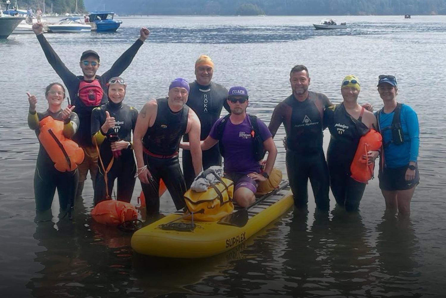 Group of swimmers with a yellow paddleboard in a body of water with mountains in the background
