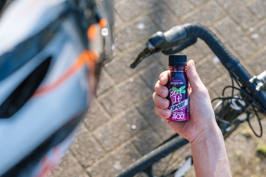 A hand holding a small bottle of purple and pink liquid supplement or energy drink, with a blurred bicycle handlebar and paved ground in the background.