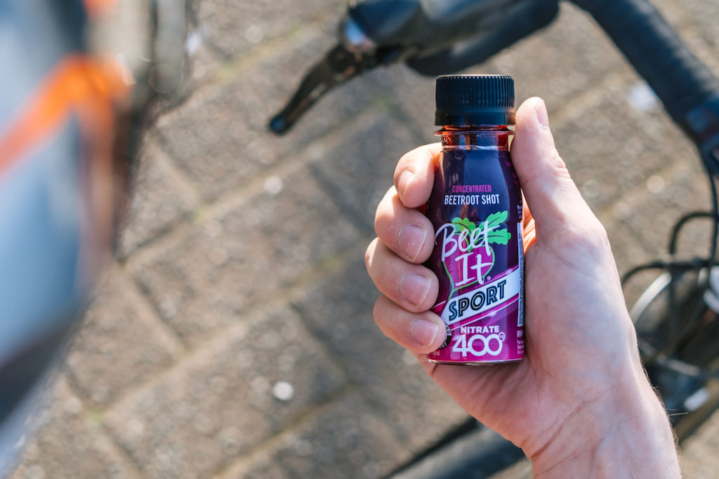 A hand holding a bottle of Beet It Sport Nitrate 400, with a blurred outdoor background showing a paved ground and bicycle handlebars.