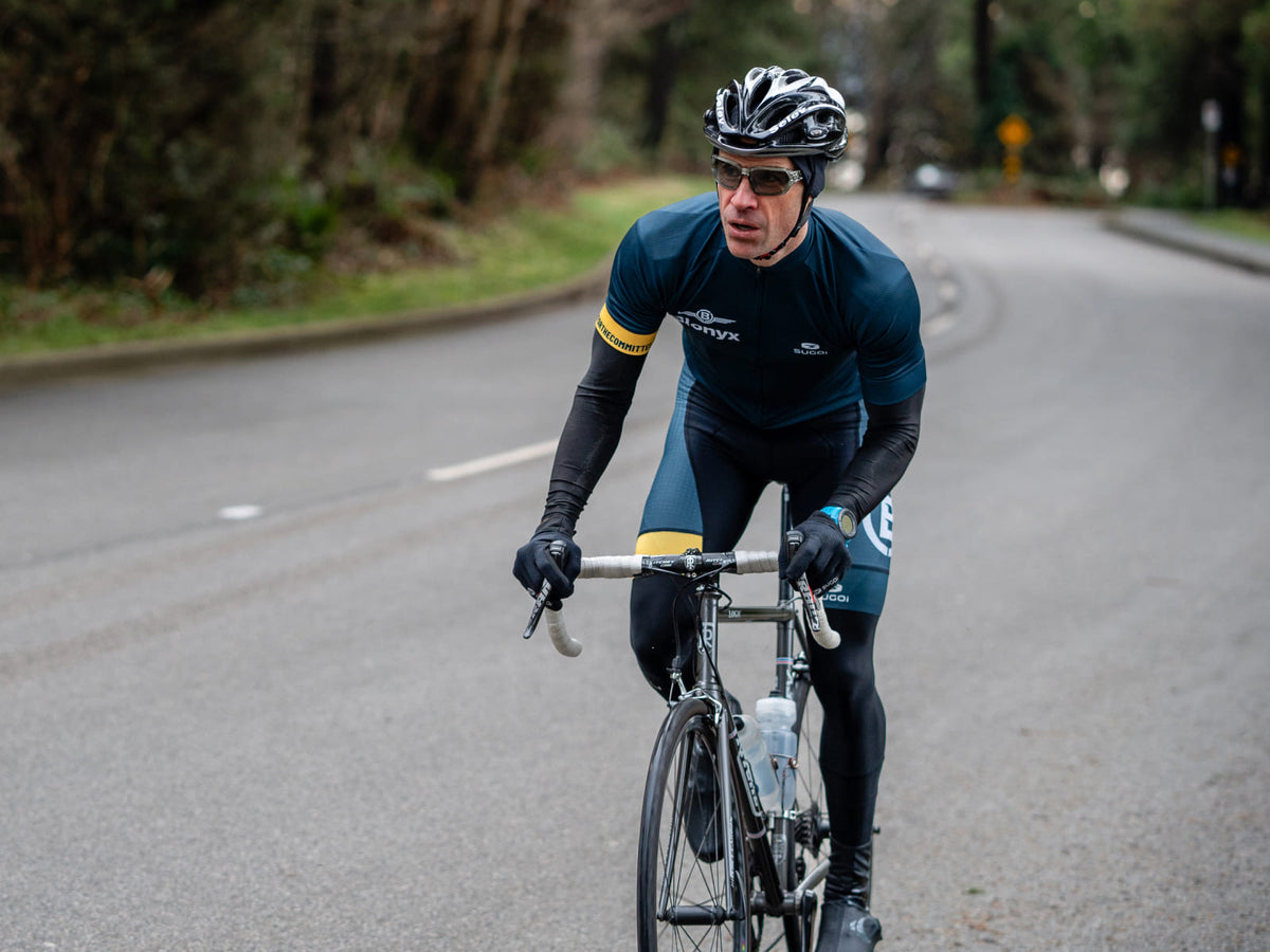 A cyclist wearing a dark navy and black cycling jersey, black tights, and a black helmet rides a road bike on a paved road with trees on both sides.