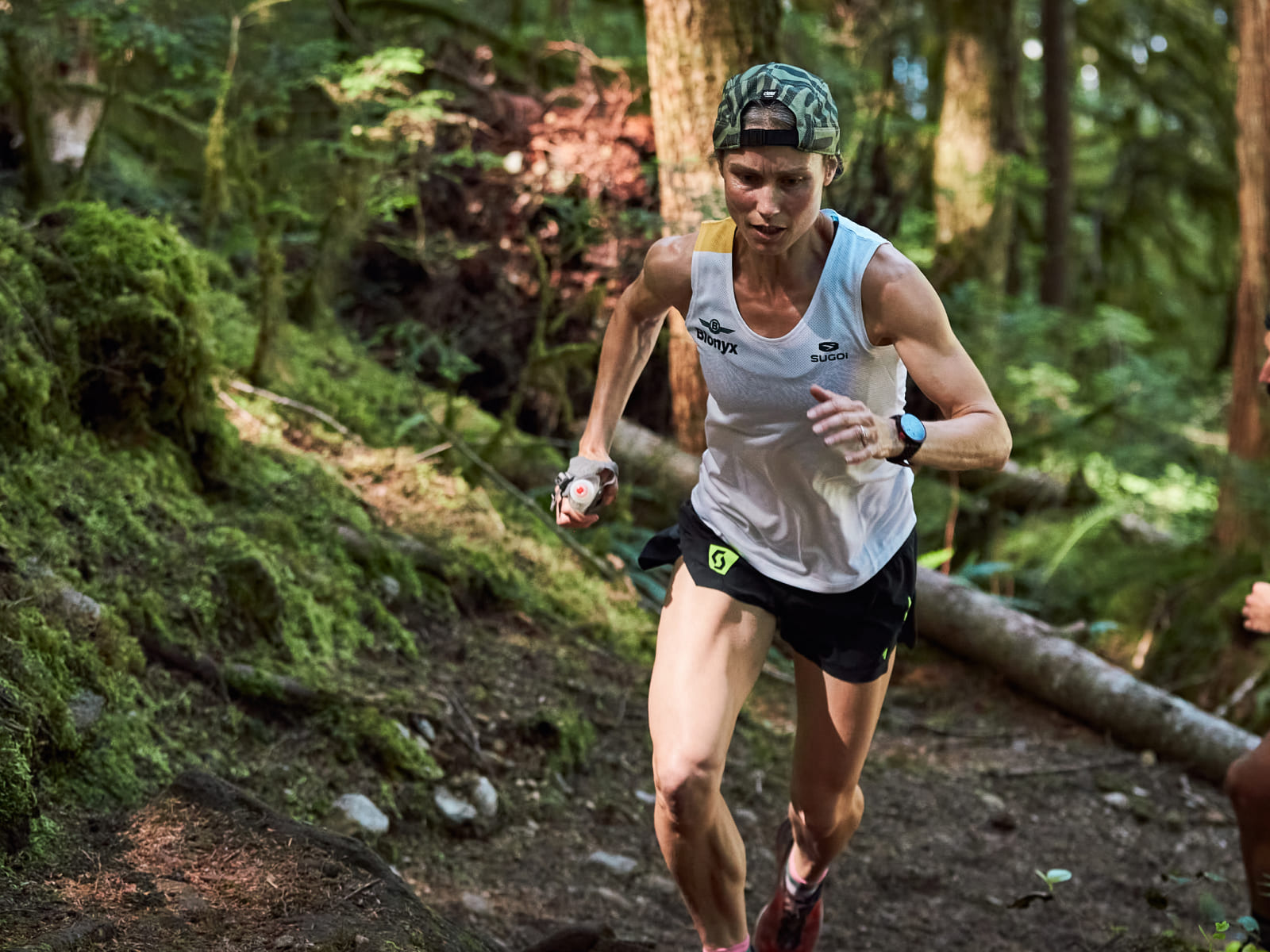 A female trail runner in a forest wearing a white sleeveless top, black shorts, and a green patterned cap, captured mid-stride on a dirt path.