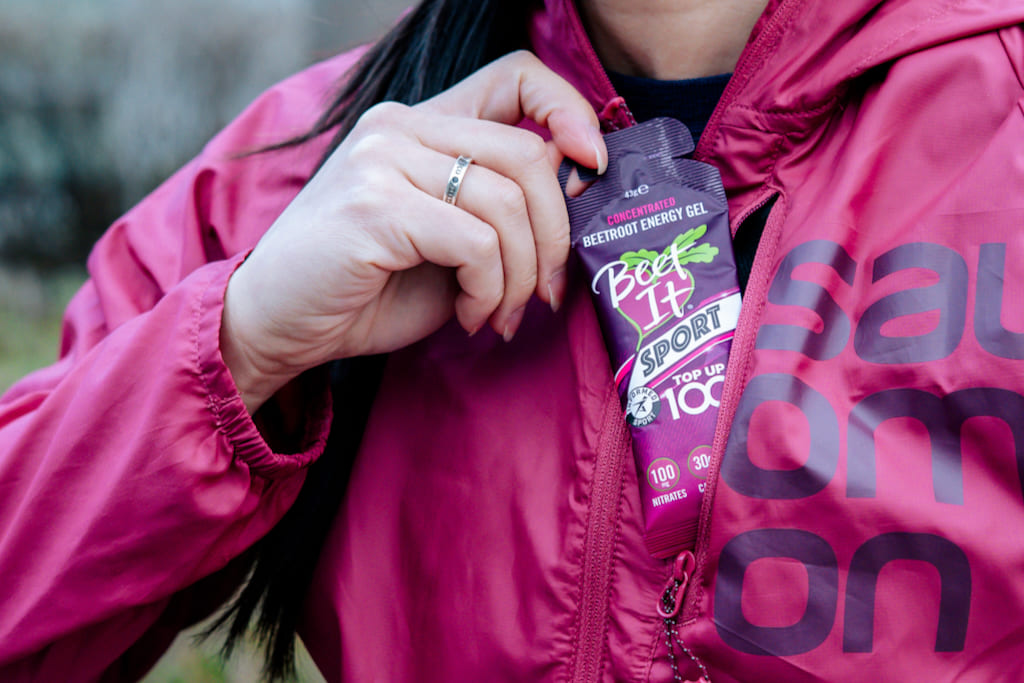 A close-up of a person wearing a bright pink jacket and pulling a purple and white Beet It Sport Top Up 100 energy gel pouch out of their pocket outdoors.