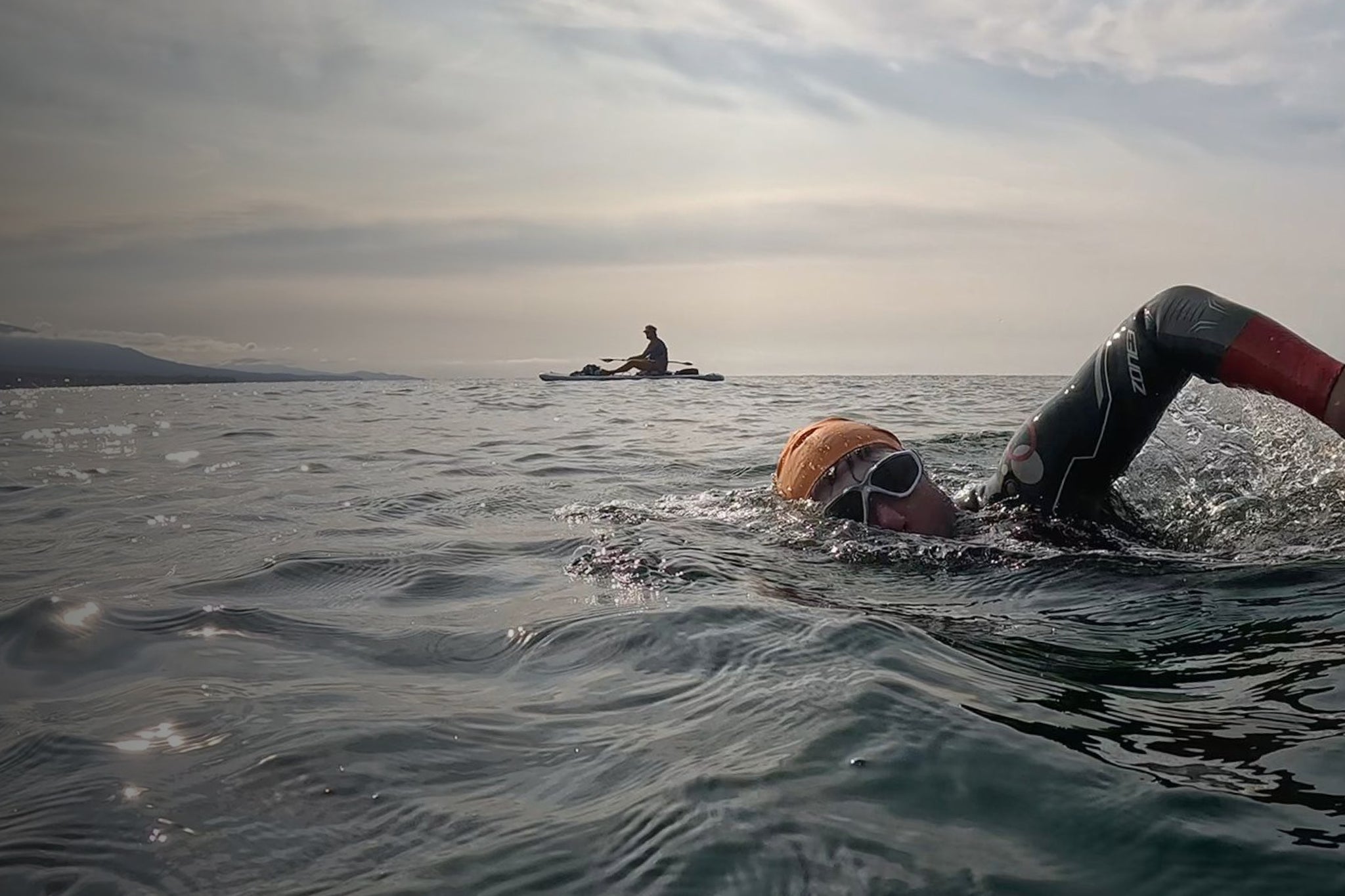 Person swimming in open water with another person in a kayak in the distance.