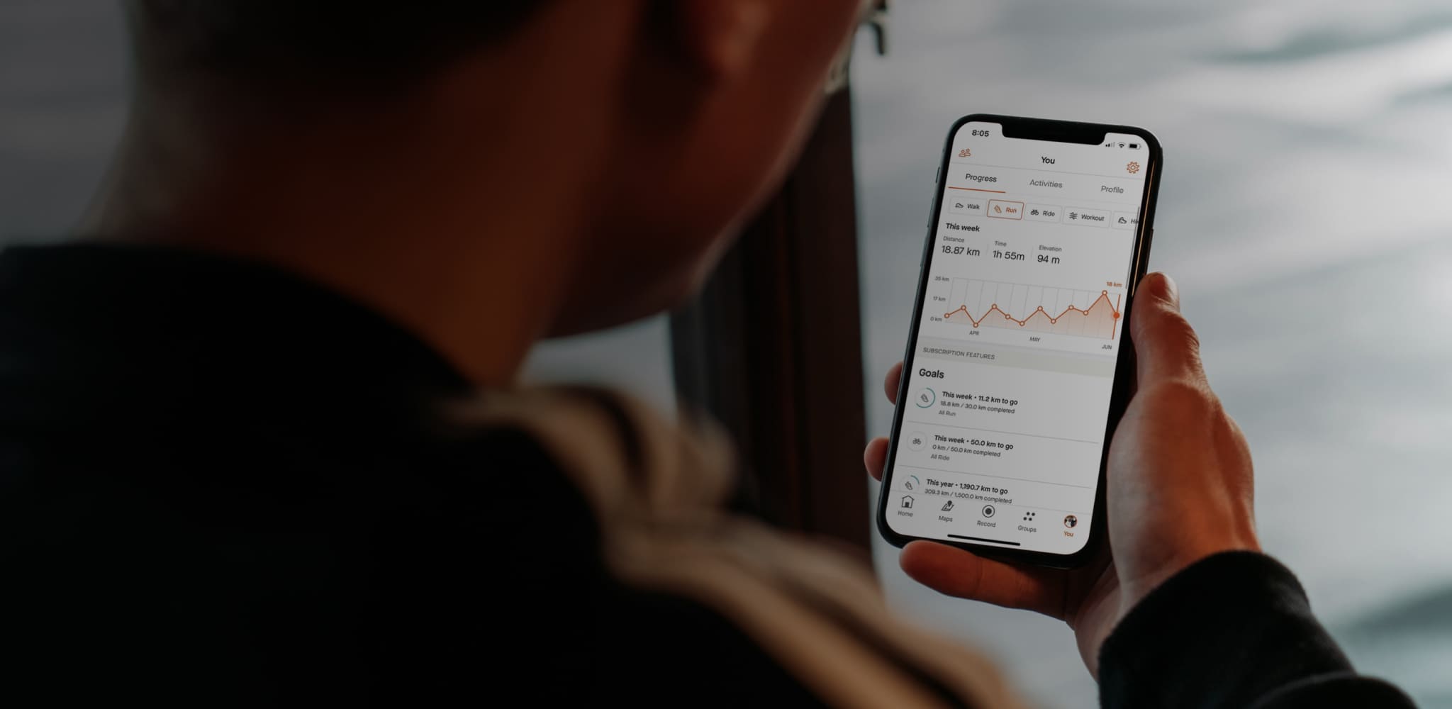 A person viewed from behind holding a smartphone with a fitness or health app screen showing a line graph, set against a soft, blurred background.
