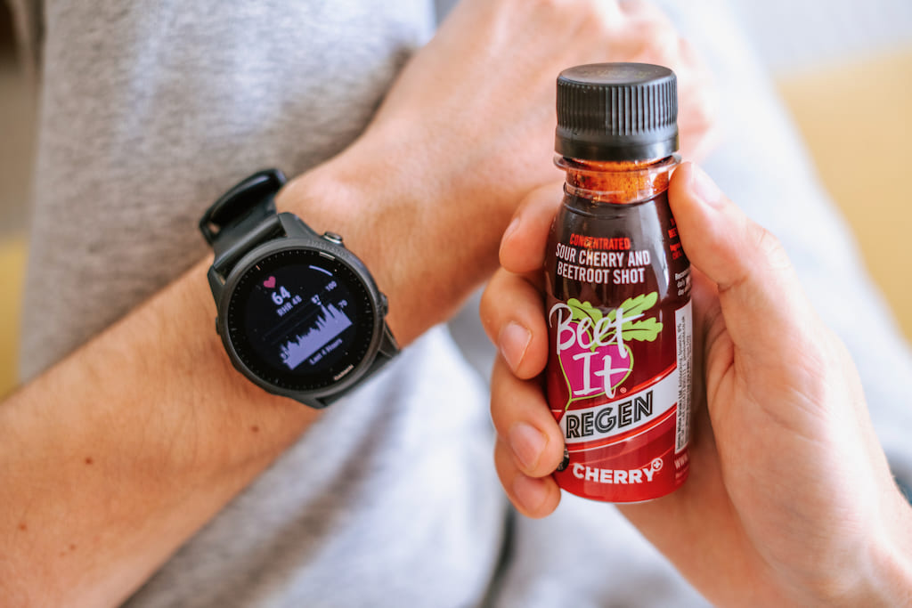 A person holds a bottle of Beet It Regen Cherry+ in one hand while wearing a smartwatch on the other wrist. The bottle label is red with black or white text, and the smartwatch screen displays a heart rate graphic.