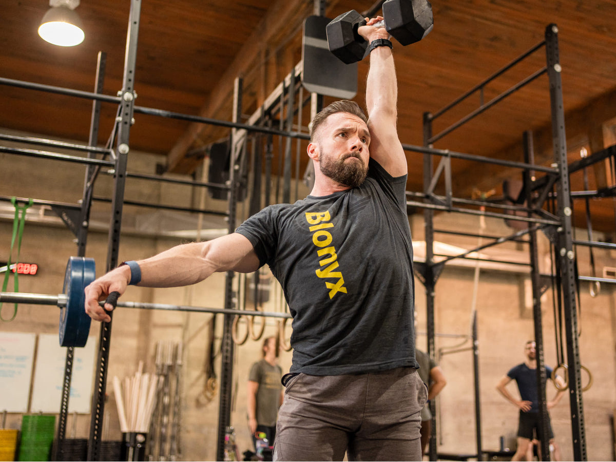 Athlete performing a single-arm dumbbell overhead lift in a functional fitness gym, wearing a black Blonyx shirt with weight racks and gym equipment in the background.