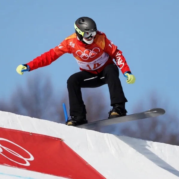 A snowboarder in a red and white Olympic-style racing jacket, black pants, and a black helmet jumps on a snowy ramp against a clear blue sky.