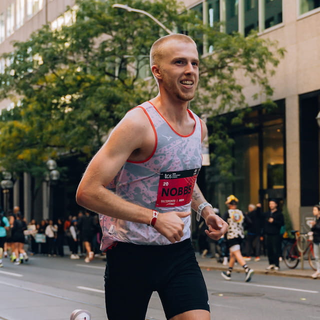 A male runner wearing a light gray patterned tank top with red trim and black shorts, running on a city street during an event. His race bib is visible, and the background shows trees, buildings, and other participants.