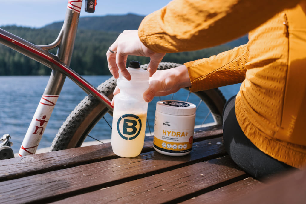 Blonyx Hydra+ real-food energy and electrolyte drink mix beside a cycling water bottle during a lakeside gravel bike ride, supporting hydration, electrolytes, and endurance performance.